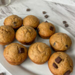 Sourdough discard banana chocolate chip muffins arranged on a white plate with chocolate chunks scattered on marble background.